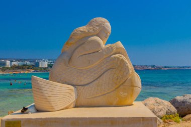 Ayia Napa, Kıbrıs - 07 Eylül 2019: Kıbrıs sahilinin önündeki heykelin gölgesinde uyuyan sokak kedisi. Sahilin arka planında inanılmaz mavi su var..