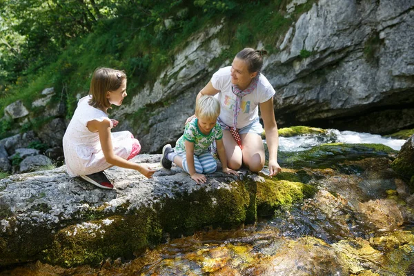 Madre con niños hablando, bebiendo agua de un arroyo de montaña puro, fresco y fresco en un viaje familiar . — Foto de Stock