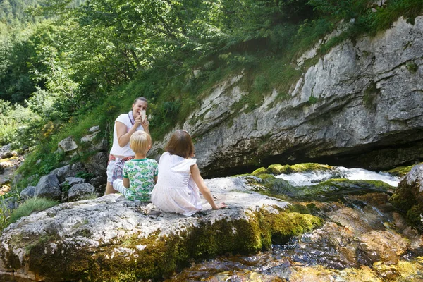 Mother taking a snapshot of kids on a family trip by a mountain stream. — Stock Photo, Image