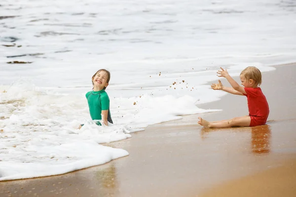 Brother and sister playing with sand and water on a tropical beach, dressed in protective wetsuit. — Stock Photo, Image