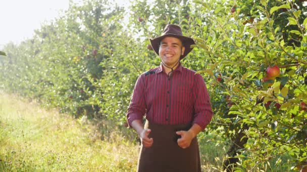帽子立っているリンゴ園で手の大きな指を出すの幸せな農家の肖像画 — ストック動画