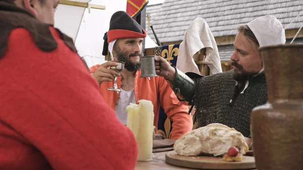 Widok z ramienia dwóch rycerzy siedzących przy stole i klinując się z filiżanką. Mężczyźni ubrani w średniowieczne ubrania. — Zdjęcie stockowe
