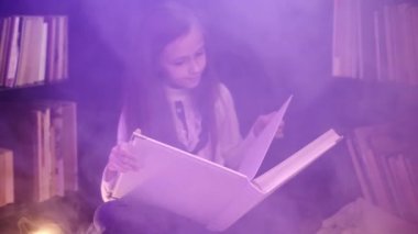 Bir kız kütüphanede bir masal kitabı açıyor, renkli dumanlar etrafında dönüyor, mumlar yakılıyor.