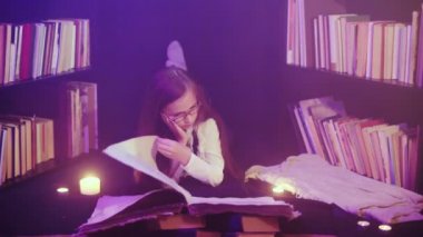 Bir kız kütüphanede bir masal kitabı açıyor, renkli dumanlar etrafında dönüyor, mumlar yakılıyor.