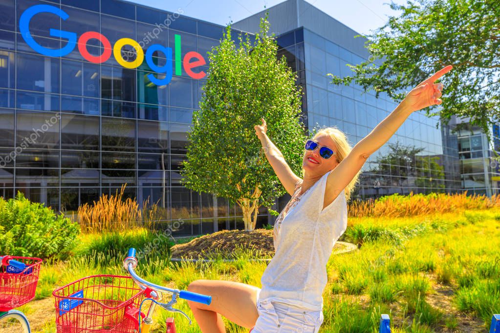 Mountain View, California, USA - August 13, 2018: Smiling tourist woman on Google bike under Google sign at Google Headquarters building. A young girl visiting the web leader company in Silicon Valley