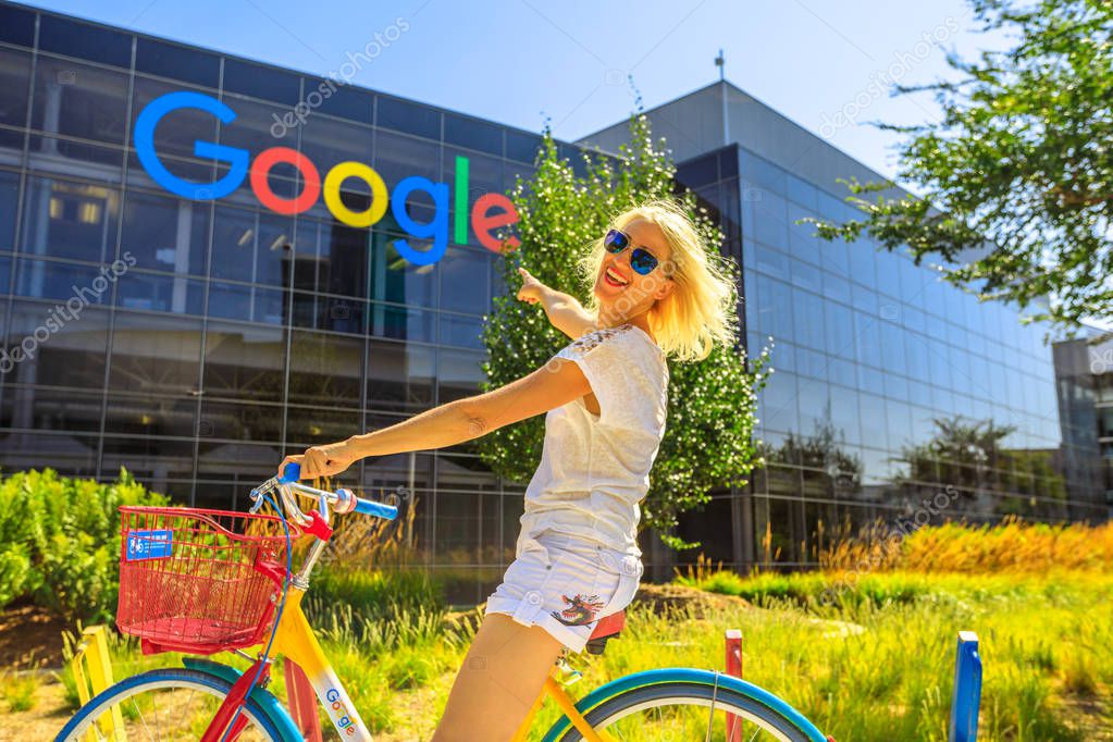 Mountain View, California, USA - August 13, 2018: Smiling woman on Google bike pointing Google sign at Googleplex Headquarters building. A young girl visiting the web leader company in Silicon Valley
