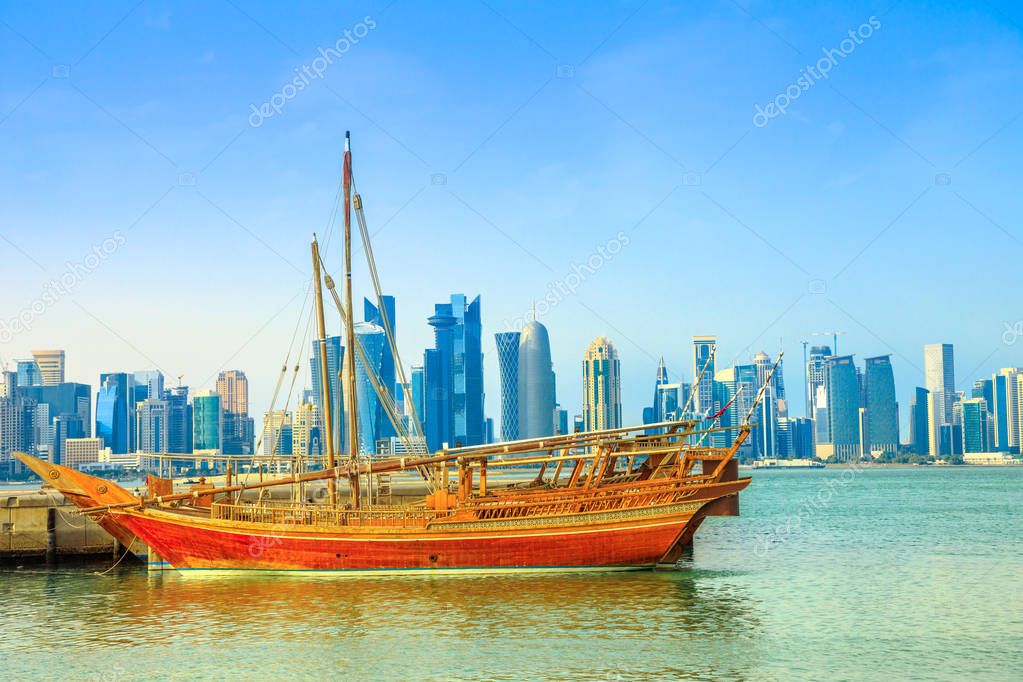 Traditional wooden dhow in foreground with seafront of Doha Bay and skyscrapers of West Bay skyline on background. Capital of Qatar, Middle East, Persian Gulf. Waterfront urban city.