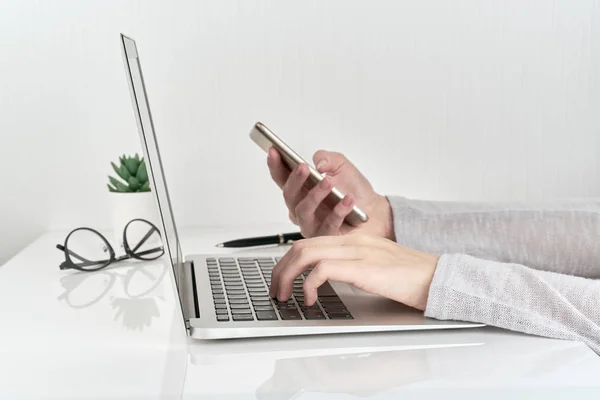 Unrecognizable woman using smartphone while typing on laptop, concept of password entry