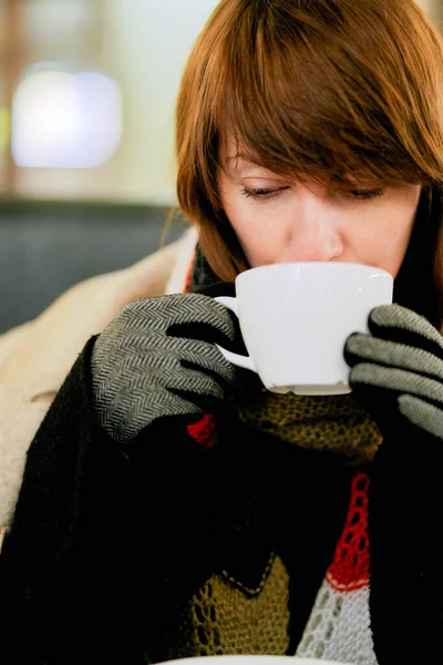 Frozen girl is warming herself with hot drink. Hands in gloves hold cup of coffee or tea