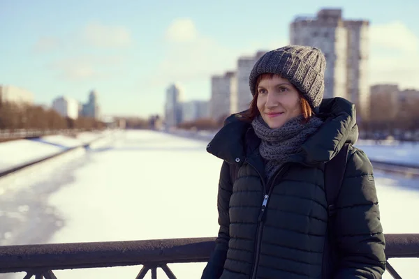 Beautiful woman stands on embankment in winter. Middle-aged female in warm hat and woolen scarf