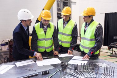 Mühendis, foreman ve emniyet kask giyen işçileri fabrika ve tartışarak kağıtları ve bluprints masada Toplantı