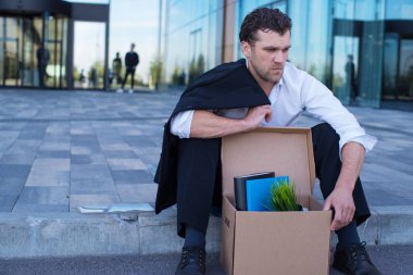 Fired business man sitting frustrated and upset on the street near office building with box of his belongings. He lost work clipart