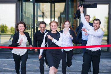 Bir grup mutlu iş adamı ofis binasından kırmızı kurdele bitiş çizgisini geçiyor.