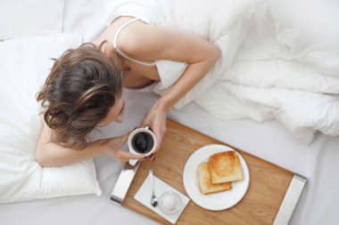 Güzel genç bir kadın yatakta kahvaltı yapıyor.