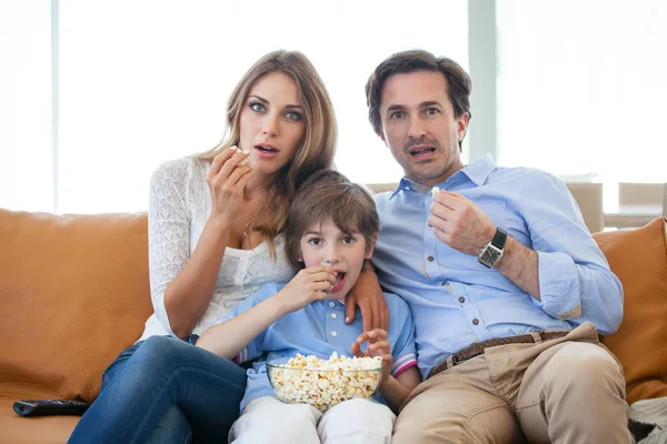年轻漂亮的父母和他们的儿子正在看电视 坐在家里的沙发上吃爆米花 — 图库照片