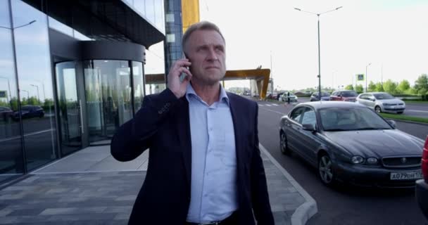 携帯電話で話しているオフィスビルに沿って野外を歩いている成熟したビジネス人 — ストック動画