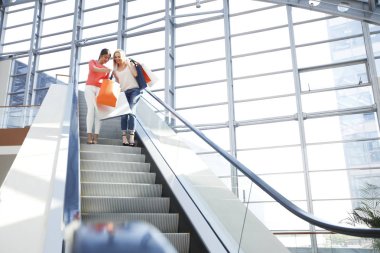 Alışveriş merkezinin yürüyen merdivenlerinde çantaya bakan güzel, mutlu kadınlar.