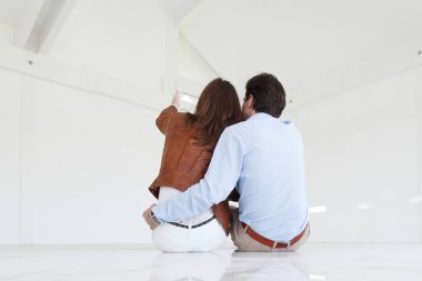 Genç çift yeni boş evin zemininde oturuyor ve gökyüzünü işaret ediyor.