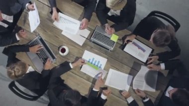 Bir grup farklı işletme yöneticisi masanın etrafında toplanarak istatistiksel veri analizlerini gösteren grafikleri tartışıyor.