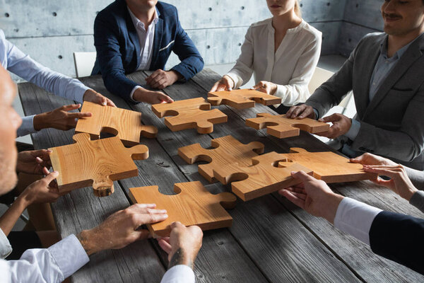 Business people team sitting around meeting table and assembling wooden jigsaw puzzle pieces unity cooperation ideas concept