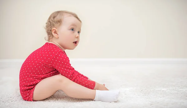 Child girl in a red T-shirt sitting on a white carpet in the room. — Stock Photo, Image