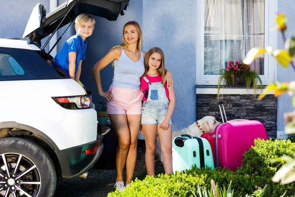 Mother, girl and boy are loading multicolored suitcases in the trunk of car. Family with a dog labrador stand near the car and ready for travel.