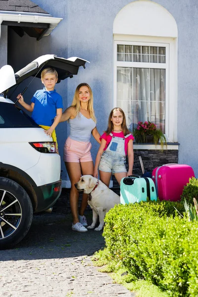 Mother, girl and boy are loading multicolored suitcases in the trunk of car. Family with a dog labrador stand near the car and ready for travel.