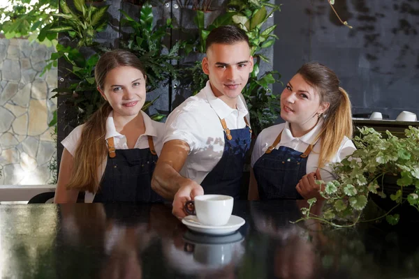 A cute young waiters in a white T-shirts and a blue aprons holding a cup of coffee in hands. Portrait of confident young male and female barista serving coffee to customer at cafe.