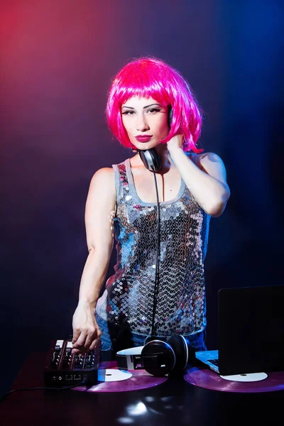 Portrait of a young woman with pink hair and vintage glasses on red and blue background. DJ woman with headphne and pink vinyls plays music disko. Glamour party on the equipment in the summer club.