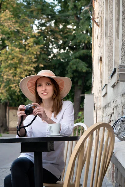 Tourist woman with vintage digital camera shooting and drinking coffee at city street cafe terrace. A young girl in a hat and a white shirt is holding a digital camera.