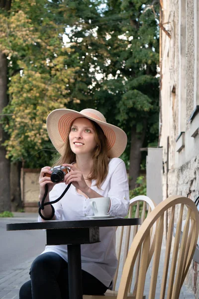 Tourist woman with vintage digital camera shooting and drinking coffee at city street cafe terrace. A young girl in a hat and a white shirt is holding a digital camera.