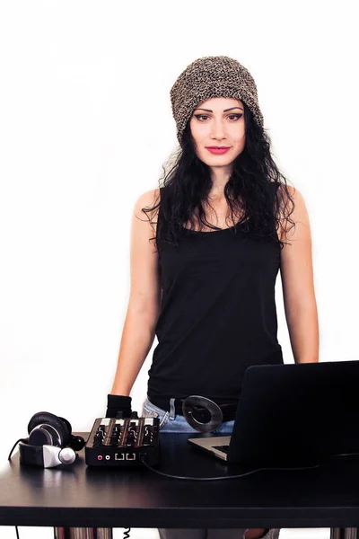 Fashion DJ woman in hat from cannabis and with headphones listening to music on the white background. Brunette in the style of casual near the table with mixer.