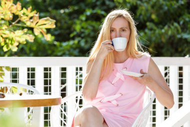 Seksi orta yaş kadın giyinmiş bir pembe elbise oturur bir yaprak arka planı. Güzel mutlu sarışın 50 artı kahve içme ve güneş zevk.