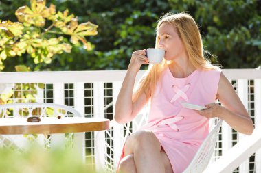 Seksi orta yaş kadın giyinmiş bir pembe elbise oturur bir yaprak arka planı. Güzel mutlu sarışın 50 artı kahve içme ve güneş zevk.