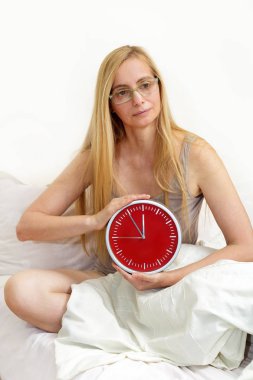 Orta 50 yaş arası artı kadın yatakta oturur ve kırmızı bir saat tutar. Bir kadının menopoz sırasında kırışıklıklar ile gerçek portre.