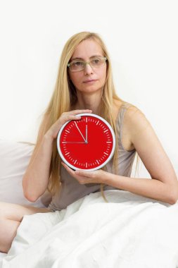 Orta 50 yaş arası artı kadın yatakta oturur ve kırmızı bir saat tutar. Bir kadının menopoz sırasında kırışıklıklar ile gerçek portre.