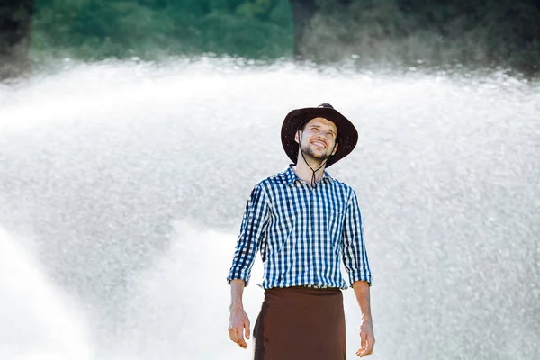 The farmer in the hat is enjoying the rain. Young man getting wet under the rain in summer.