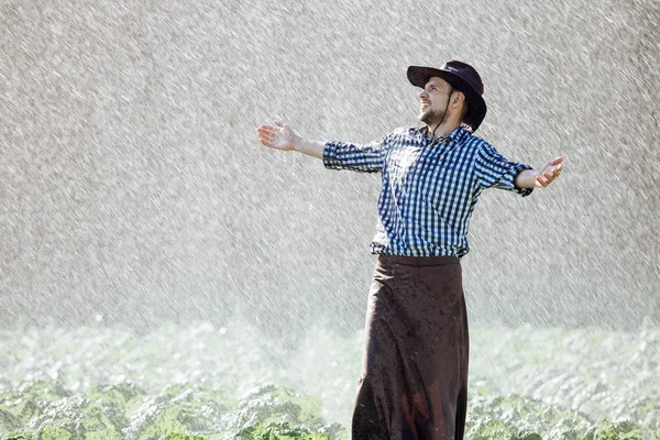The farmer in the hat is enjoying the rain. Young man getting wet under the rain in summer.