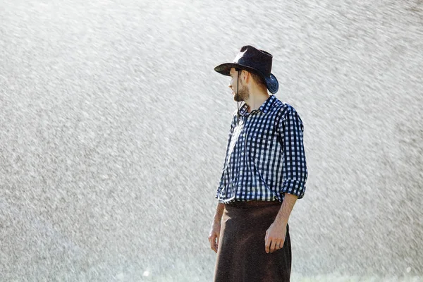 The farmer in the hat is enjoying the rain. Young man getting wet under the rain in summer.