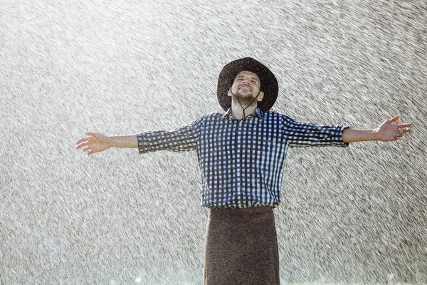 The farmer in the hat is enjoying the rain. Young man getting wet under the rain in summer.