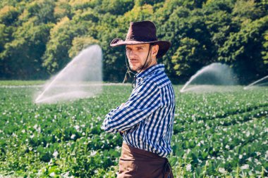 Çiftçi sahada sulama veya yağmurlama sistemi arka plan üzerinde duruyor. Genç bir ziraat mühendisi bir plantasyonda olduğunu ve hasat kalite denetimleri.