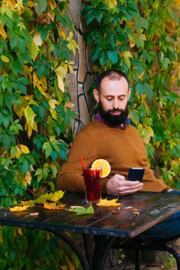 Sakallı adam sonbahar yaprakları arka plan üzerinde bir kafede oturuyor. Esmer sarı kazaklı içecekler ve sıcak mulled şarap bir kafe ya da bahçe içinde olsun.