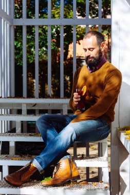 Sakallı adam beyaz merdiven içme üzerinde mulled şarap ve sonbahar güneş ışığı tadını çıkarın. İşadamı ofiste çalışıyor, bir cam bir kırmızı sıcak içecekle tutar.