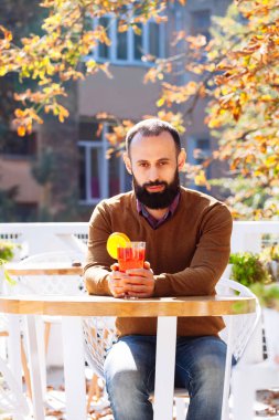 Sakallı adam sonbahar yaprakları arka plan üzerinde bir kafede oturuyor. Esmer sarı kazaklı içecekler ve sıcak mulled şarap bir kafe ya da bahçe içinde olsun.