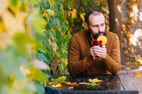 Beard man is sitting in a cafe on the background of autumn leaves. Brunette in yellow sweater drinks and get warm mulled wine in a cafe or garden.