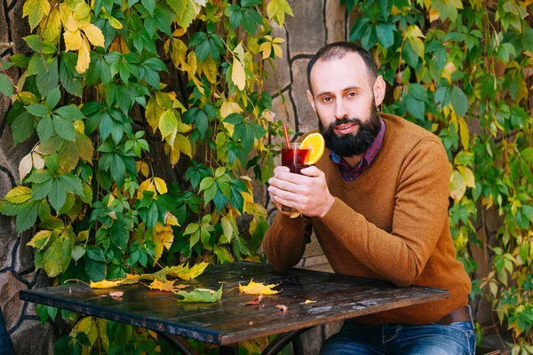 Beard man is sitting in a cafe on the background of autumn leaves. Brunette in yellow sweater drinks and get warm mulled wine in a cafe or garden.