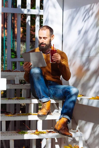 Bearded man in a cafe drinking mulled wine. Businessman works in office, holds a glass with a red hot drink and uses tablet.