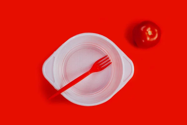 White plastic plate, tomato and red fork on red background.