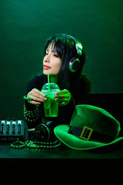Portrait of DJ young irish girl on green background. DJ woman with headphne and green hat plays music on the party of St. Patrick.
