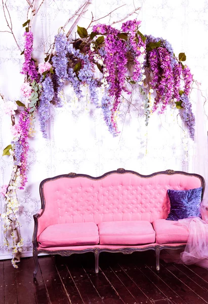 Old pink sofa with blue pillow on a white wall decorated with pink and purple flowers.